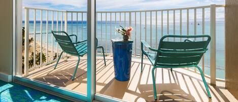 A balcony with two green chairs and a vase of flowers, overlooking a beach and the ocean.