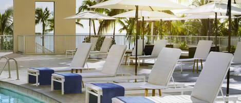 A poolside area with lounge chairs and umbrellas, a pool, and palm trees in the background.