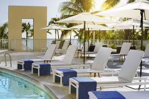 Un área junto a la piscina con sillones de descanso y sombrillas, una piscina y palmeras al fondo.