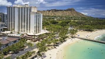A high-rise hotel with a pool and beach access, surrounded by palm trees and a mountain in the background.