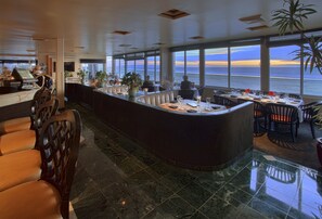 Un amplio bar con una barra curva, distribución de asientos y grandes ventanales con vistas al océano.