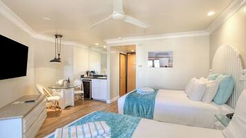 A hotel room with two beds, a desk, a chair, a small table, a ceiling fan, and a view of the kitchen and bathroom.