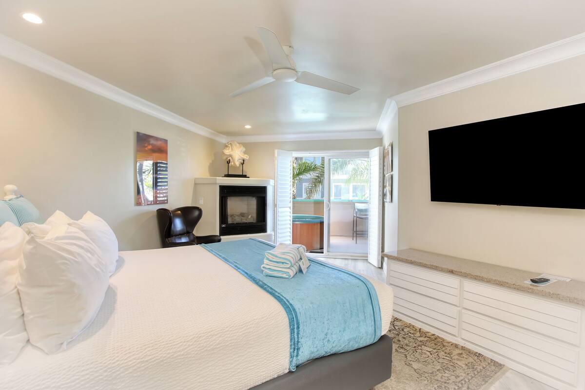 A bedroom with a bed, a ceiling fan, a fireplace, a TV, and a view of the outside.