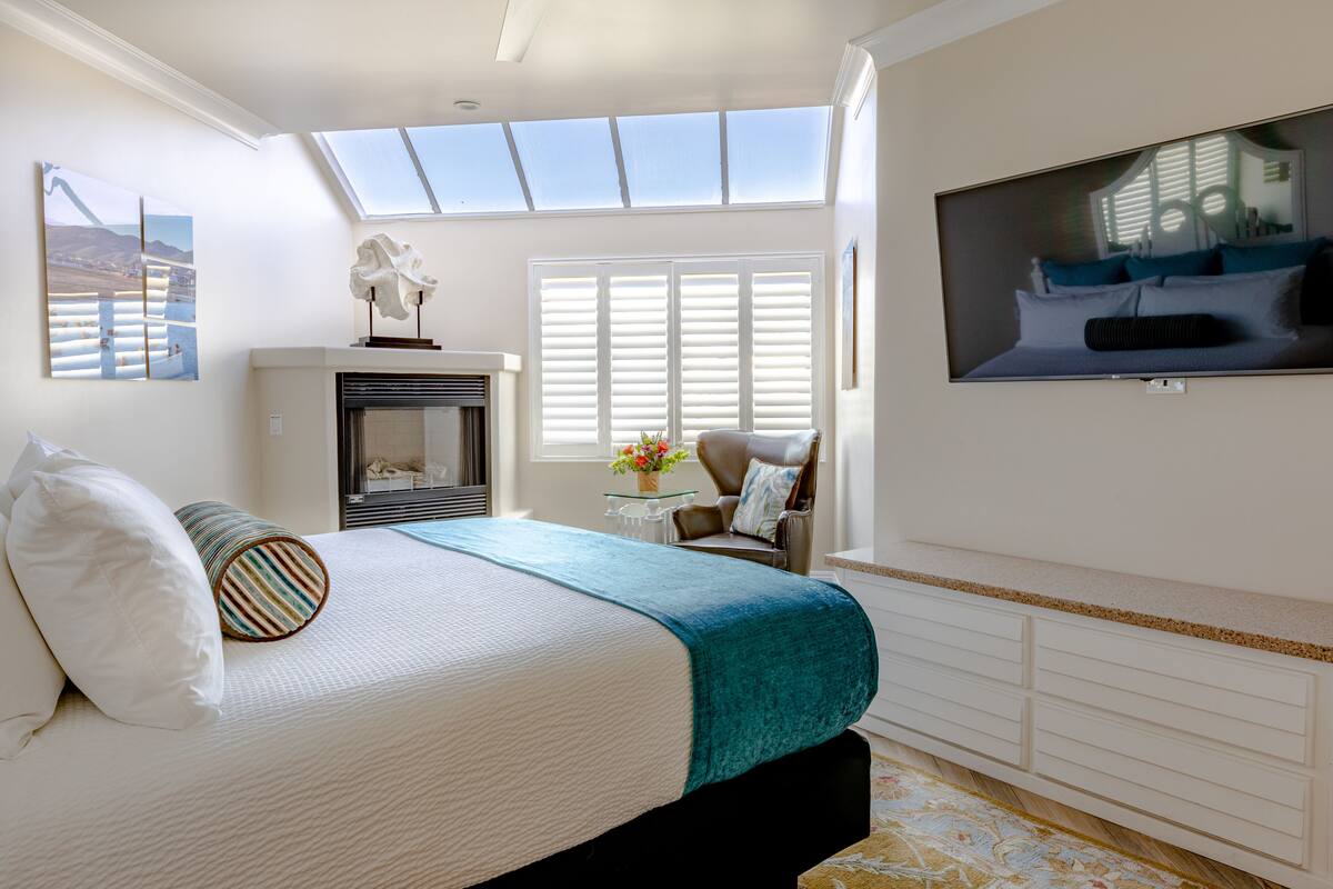 A bedroom with a bed, a television mounted on the wall, a fireplace, and a skylight.