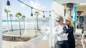 A couple standing by a glass balustrade overlooking a beach with palm trees and a pier. - SeaVenture Beach Hotel (Pismo Beach)