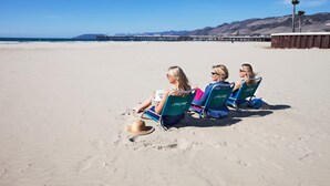 Three people sitting on beach chairs facing the ocean, with a wooden pier and mountains in the background. - SeaVenture Beach Hotel (Pismo Beach)