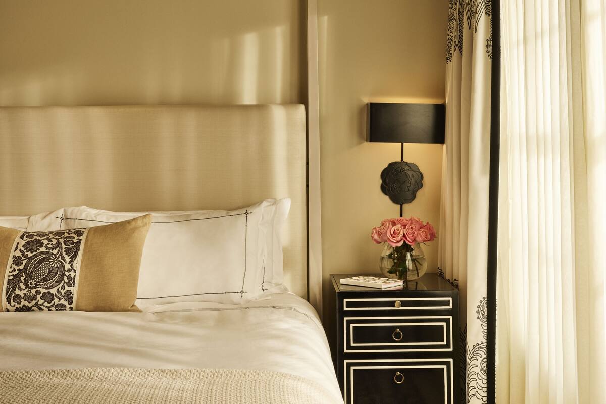 A neatly made bed with white linens and decorative pillows, a bedside table with a lamp and a vase of flowers, and a window with sheer curtains.