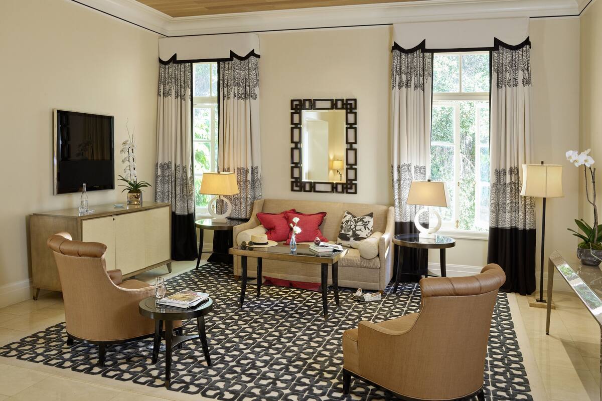 A well-lit living room with a sofa, two armchairs, a coffee table, and a TV. There are two windows with curtains, a mirror, and a lamp on a side table.