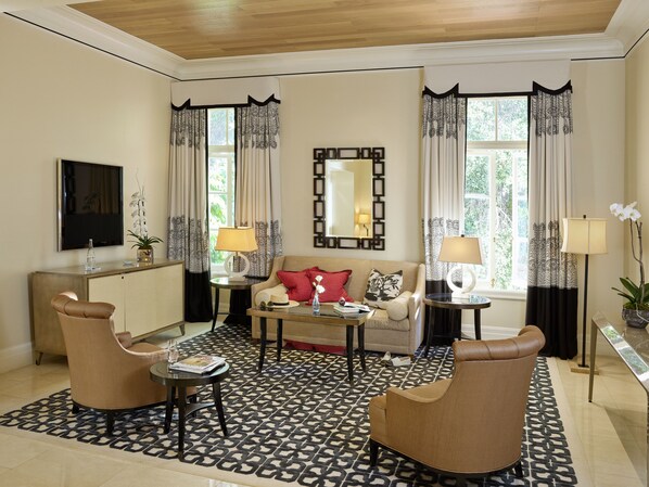 A well-lit living room with a sofa, two armchairs, a coffee table, and a TV. There are two windows with curtains, a mirror, and a lamp on a side table.