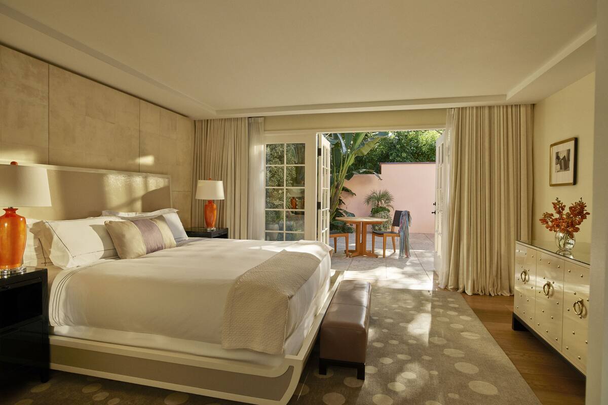 A hotel room with a bed, bedside table, lamp, bench, and a view of an outdoor patio with a table and chairs.