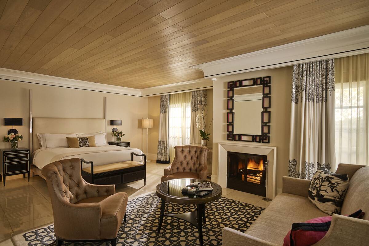 A hotel room with a bed, a sitting area with a sofa and armchair, a fireplace, and a television on the wall.