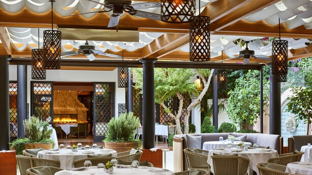 An outdoor dining area with wicker chairs and round tables, covered by a wooden pergola with hanging lights. - Hotel Bel-Air - Dorchester Collection (Los Angeles)