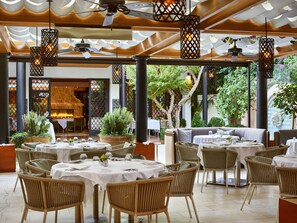 An outdoor dining area with wicker chairs and round tables, covered by a wooden pergola with hanging lights. - Hotel Bel-Air - Dorchester Collection (Los Angeles)