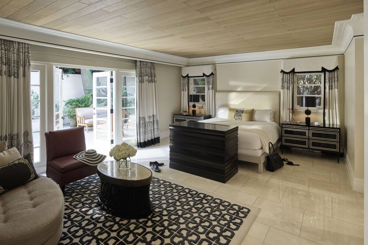 A spacious bedroom with a large bed, a dresser, a desk, and a chair. There is a window with curtains, a door leading to a balcony, and a patterned rug on the floor.
