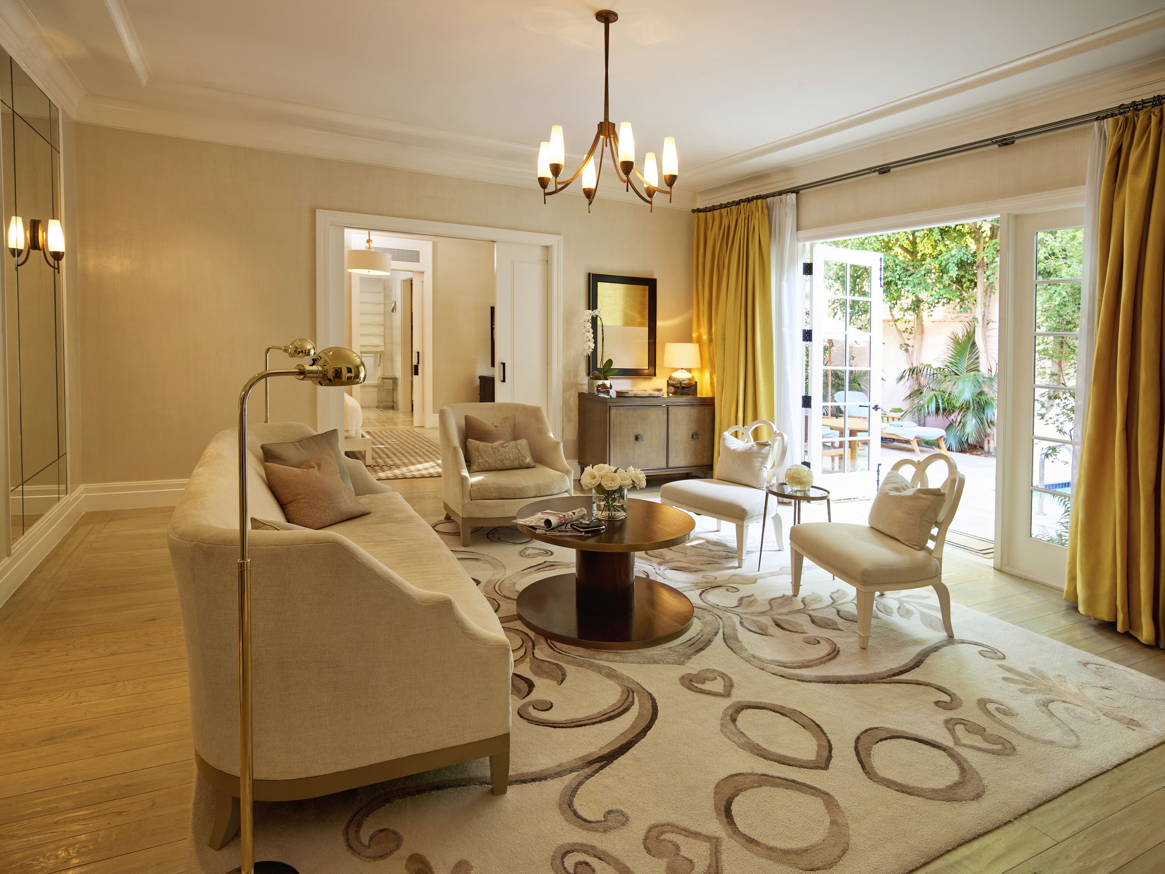 A spacious living room with a beige sofa, armchairs, a coffee table, and a chandelier. There is a large window with curtains and a view of greenery outside.