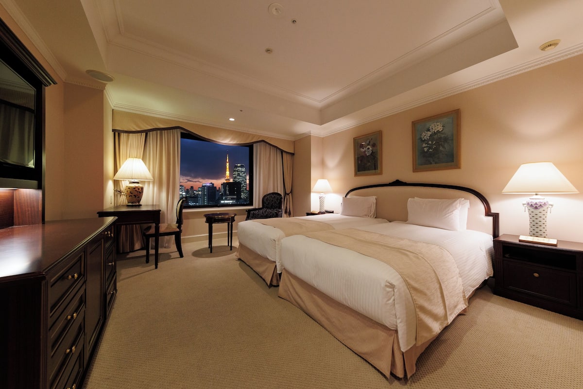 A hotel room with two beds, a desk, a chair, and a view of a cityscape at night.