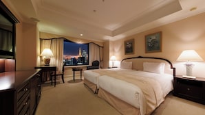 A hotel room with two beds, a desk, a chair, and a view of a cityscape at night.