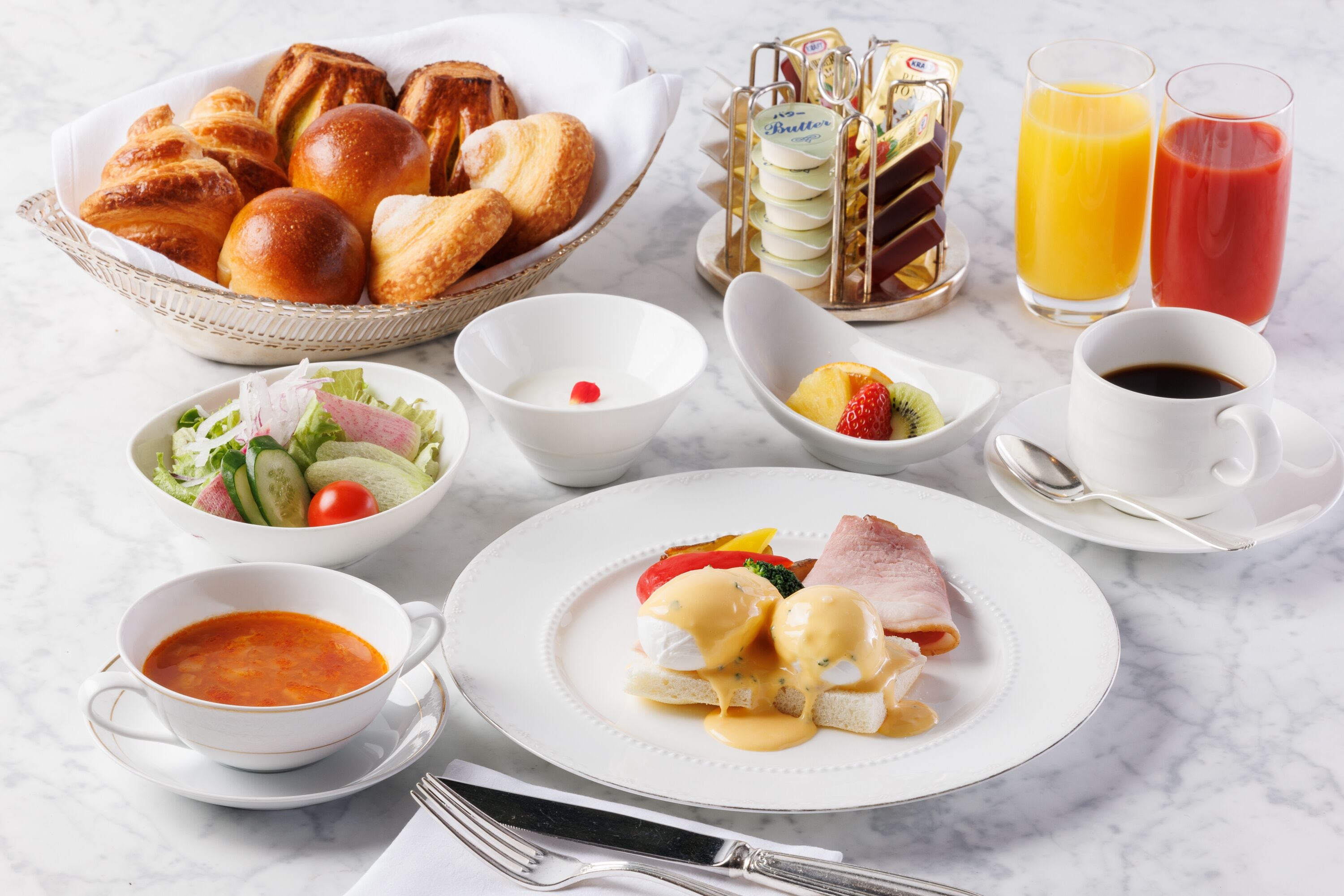 A breakfast setting with croissants, a fruit bowl, a plate of eggs, a bowl of soup, and a glass of orange juice.