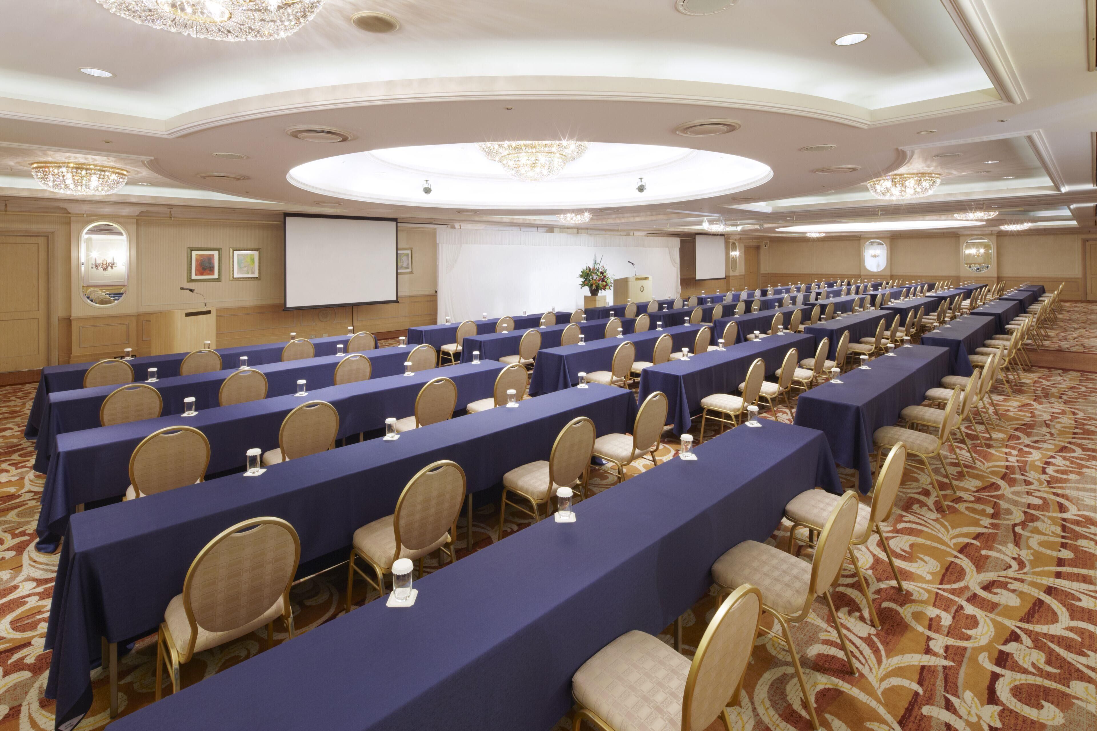 A conference room with rows of tables and chairs, a projector screen, and a carpeted floor.