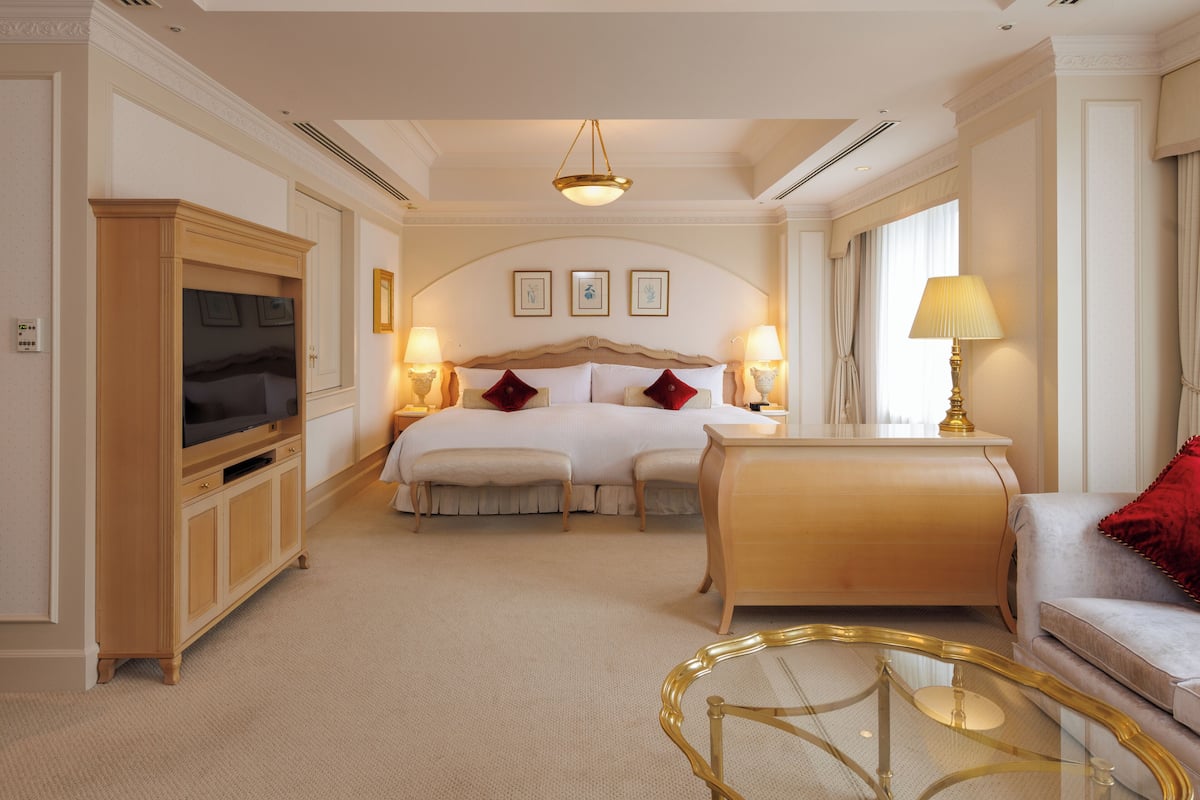 A hotel room with a bed, bedside lamps, a television, a sofa, and a glass coffee table.