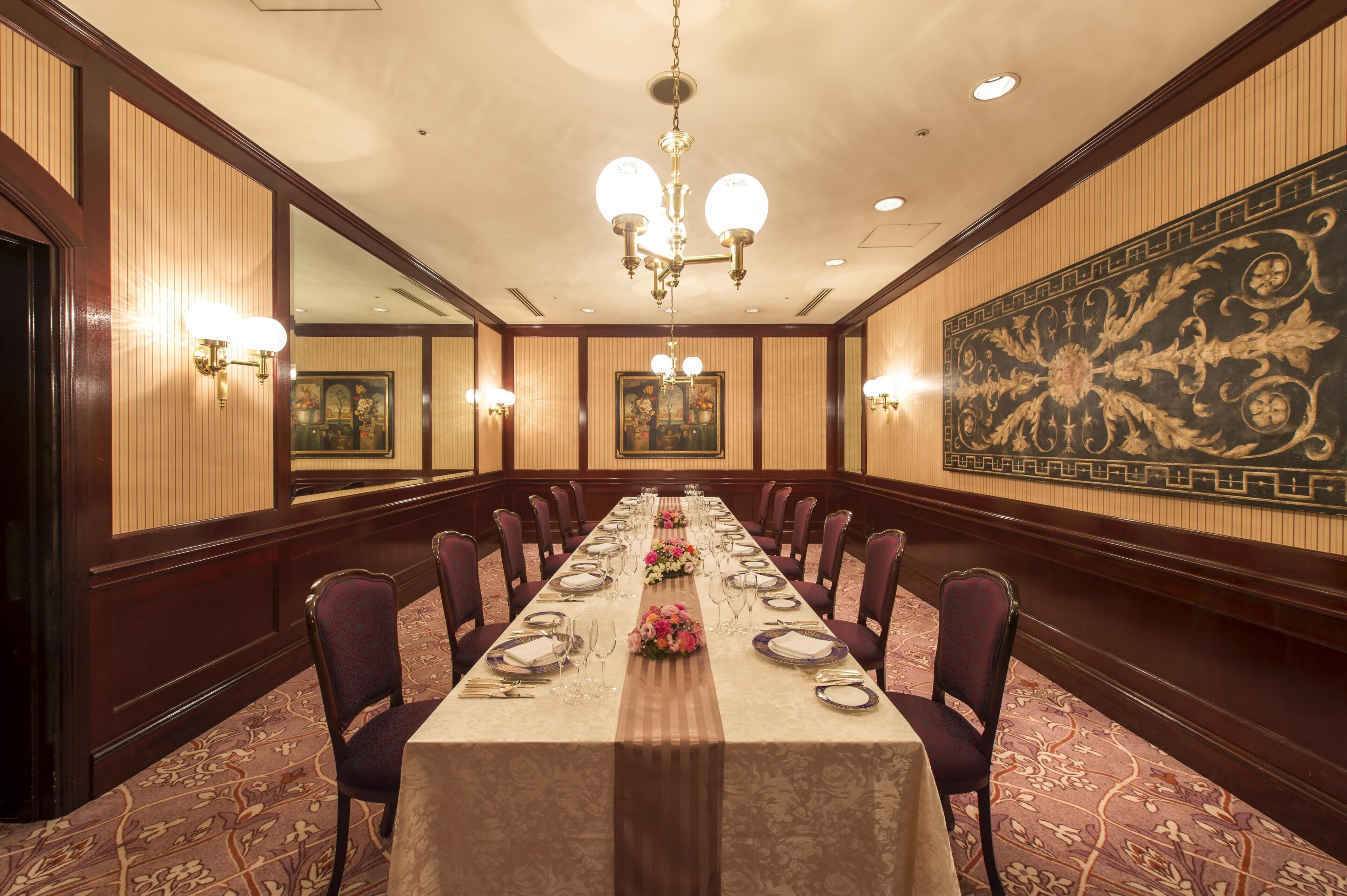 Una larga mesa de banquete dispuesta para un evento formal, con sillas a ambos lados, un candelabro y luces en la pared.