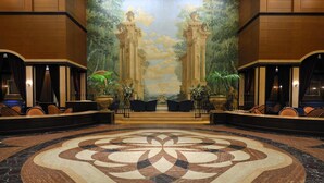 A lobby with a large floral carpet, a mural with columns, and seating areas with plants.