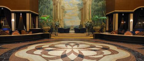 Un hall con un gran tapiz floral, un mural con columnas y zonas de estar con plantas.