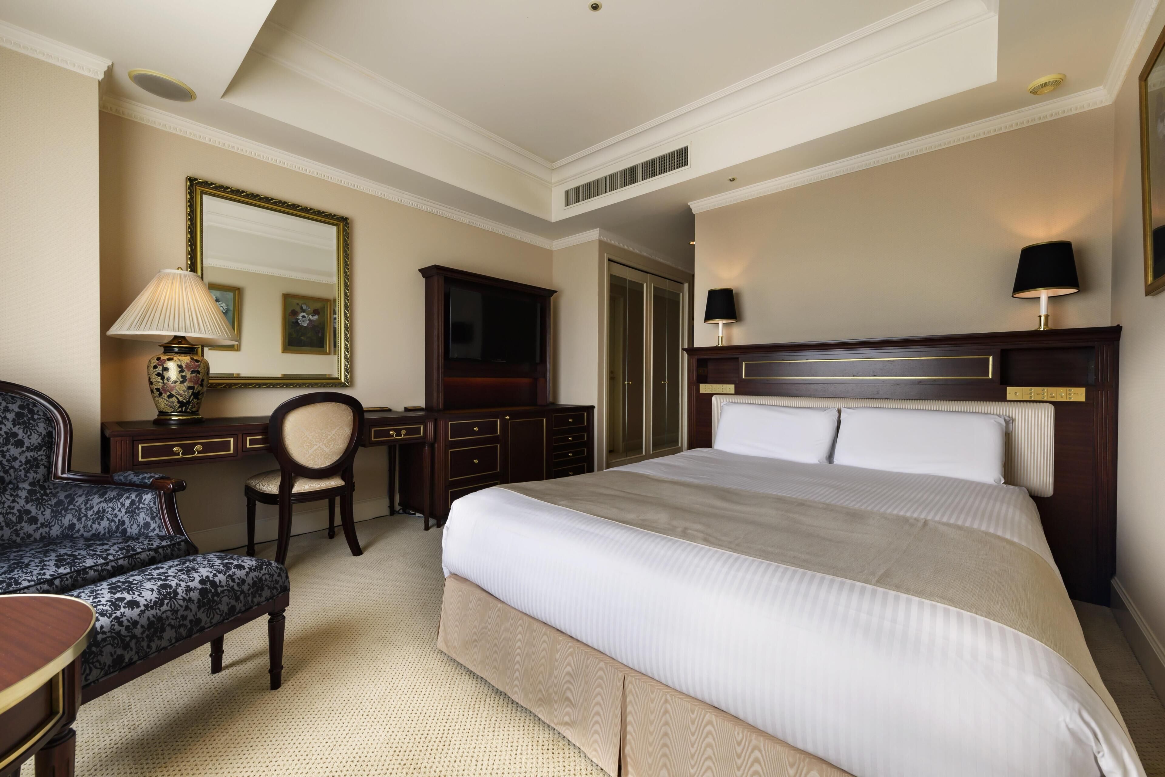 A hotel room with a large bed, a desk with a chair, a dresser, a mirror, and a lamp.