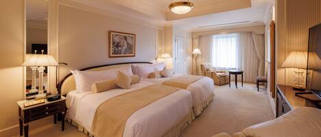 Une chambre d’hôtel avec deux grands lits, un coin salon comprenant un fauteuil et une petite table, un grand miroir et un téléviseur.