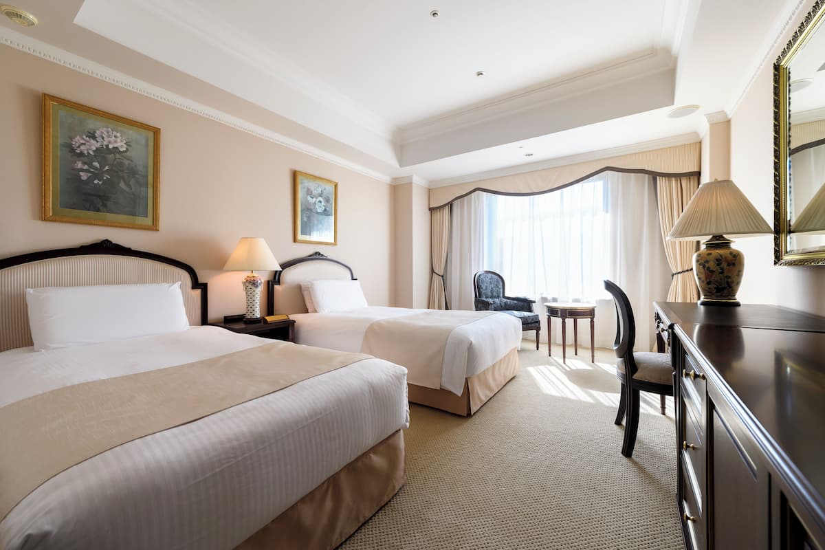 A hotel room with two beds, a desk, a chair, and a lamp.