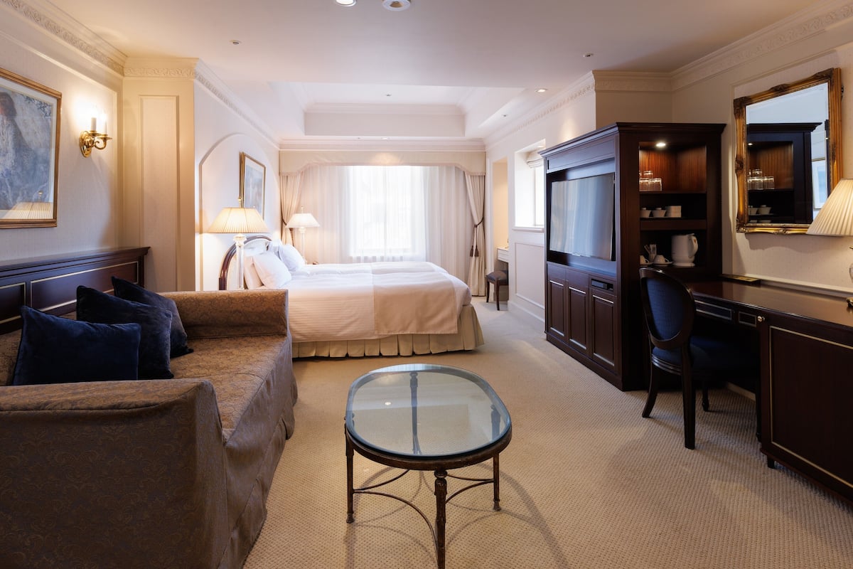 A hotel room with a bed, a sofa, a glass-top table, a TV, and a desk with a chair.