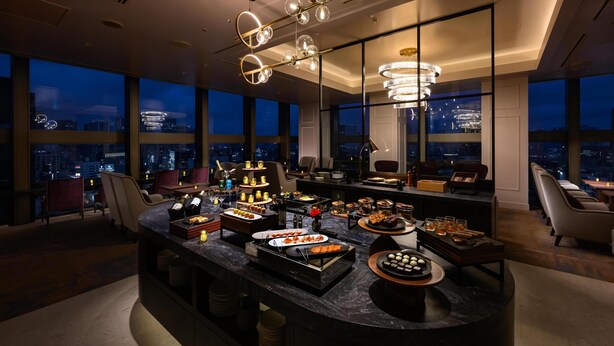 A modern bar area with a marble countertop, various food and drink options, and a city view through large windows.