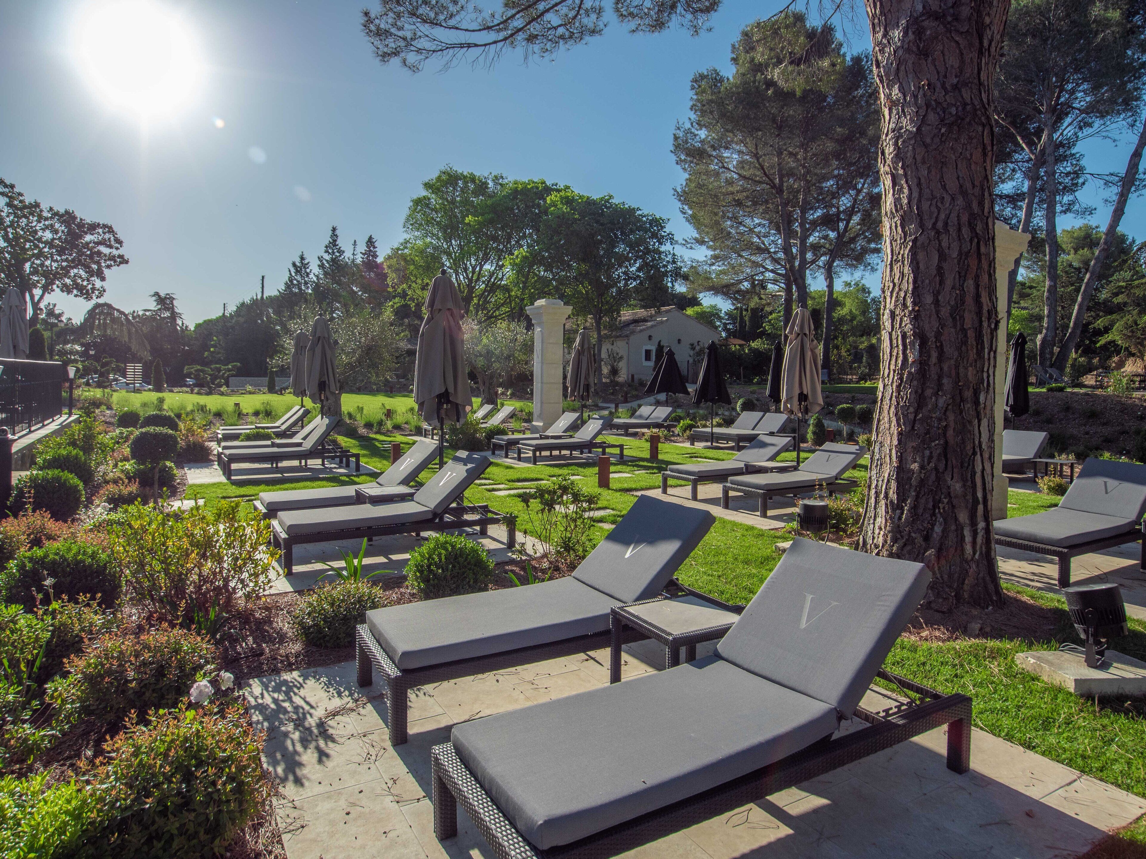 A sunlit outdoor lounge area with multiple lounge chairs and umbrellas, surrounded by well-maintained greenery and trees.