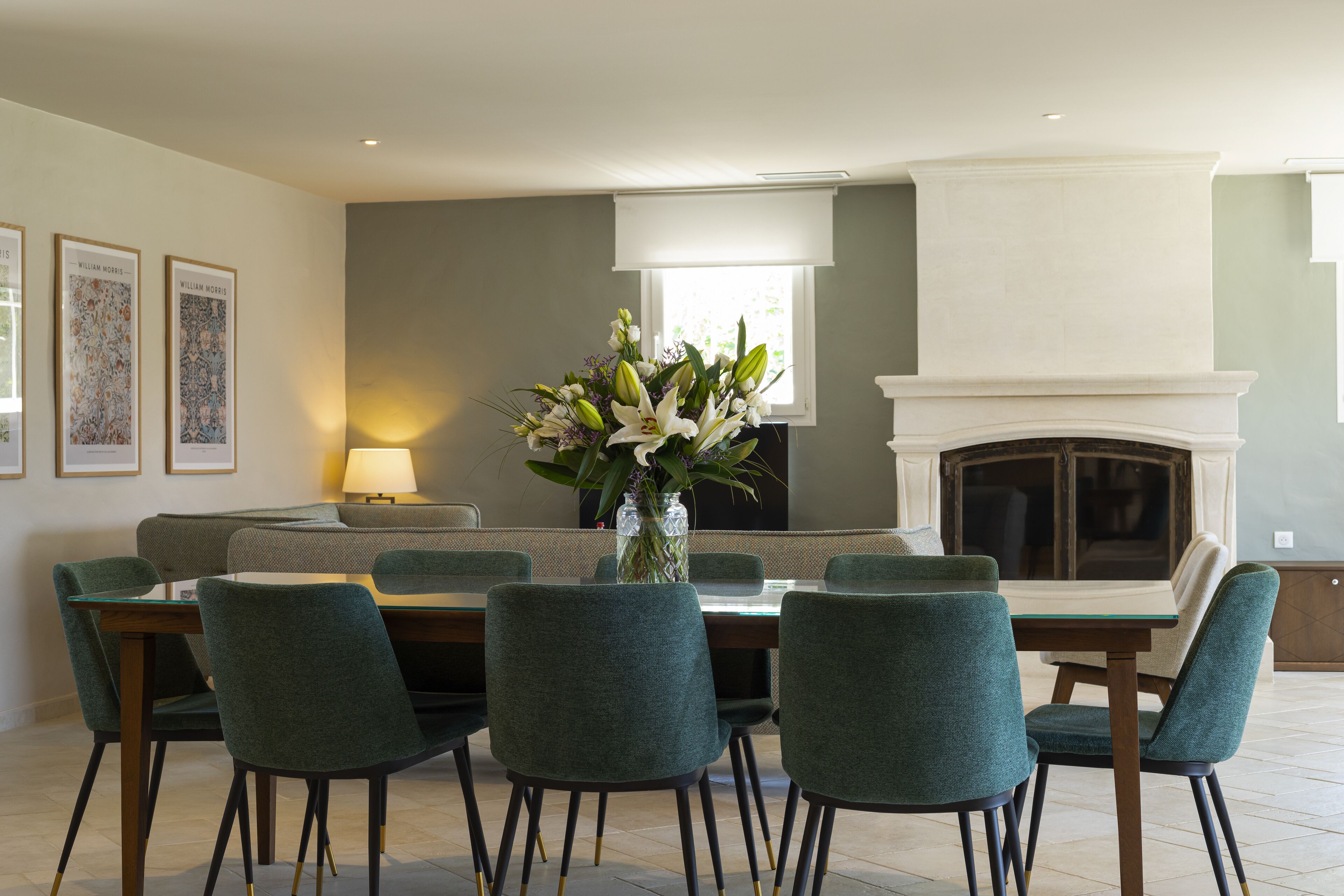 A dining area with a table set for six, surrounded by six teal chairs, a vase with flowers, and a fireplace in the background.