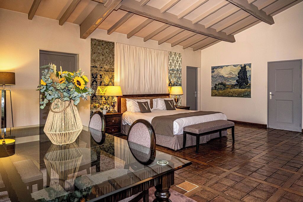 A well-lit bedroom with a bed, bedside tables, a vase of flowers, and a painting on the wall.