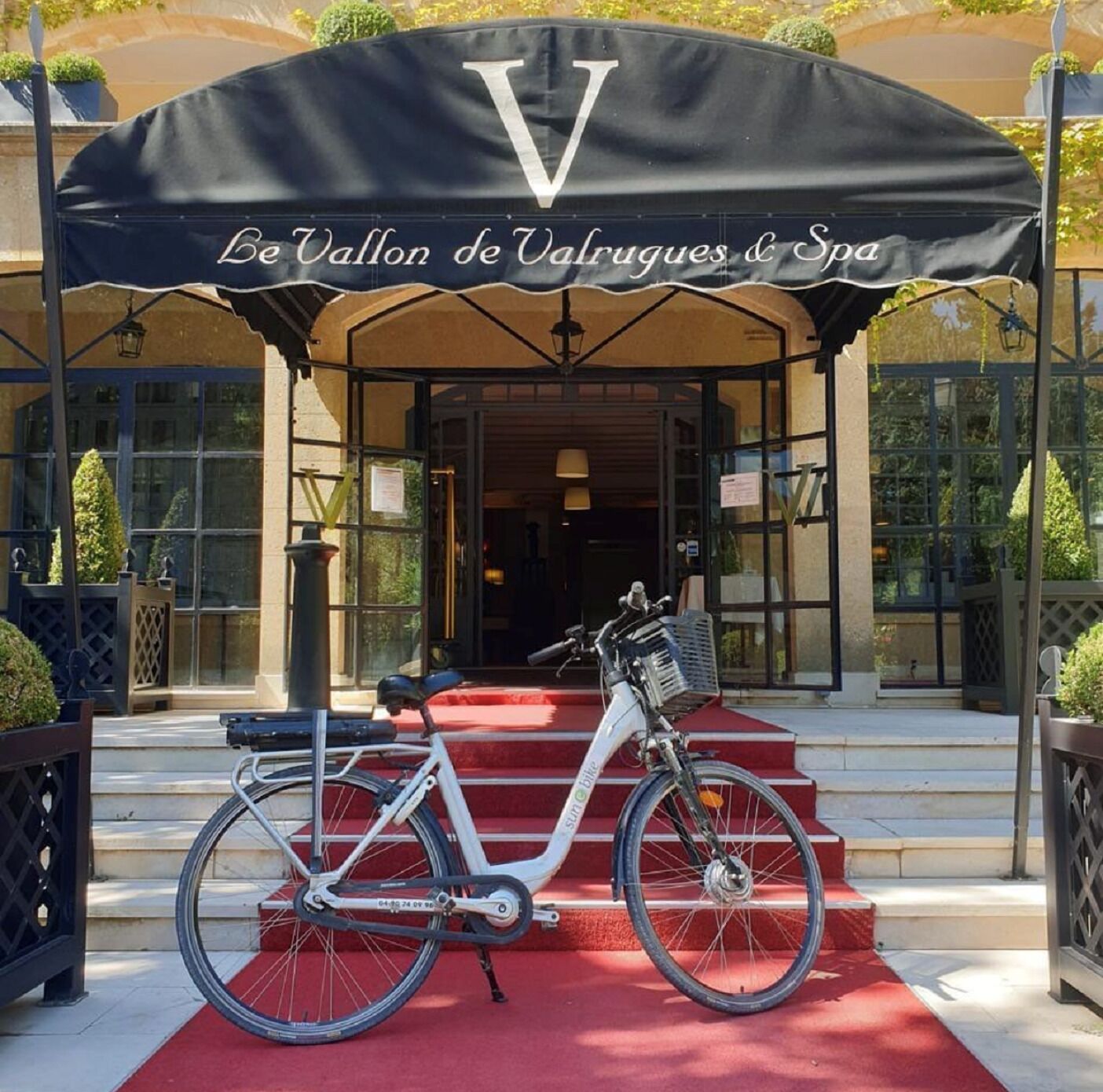 A bicycle parked on a red carpet in front of a building with a large awning displaying "Le Vallon de Valruges & Spa".