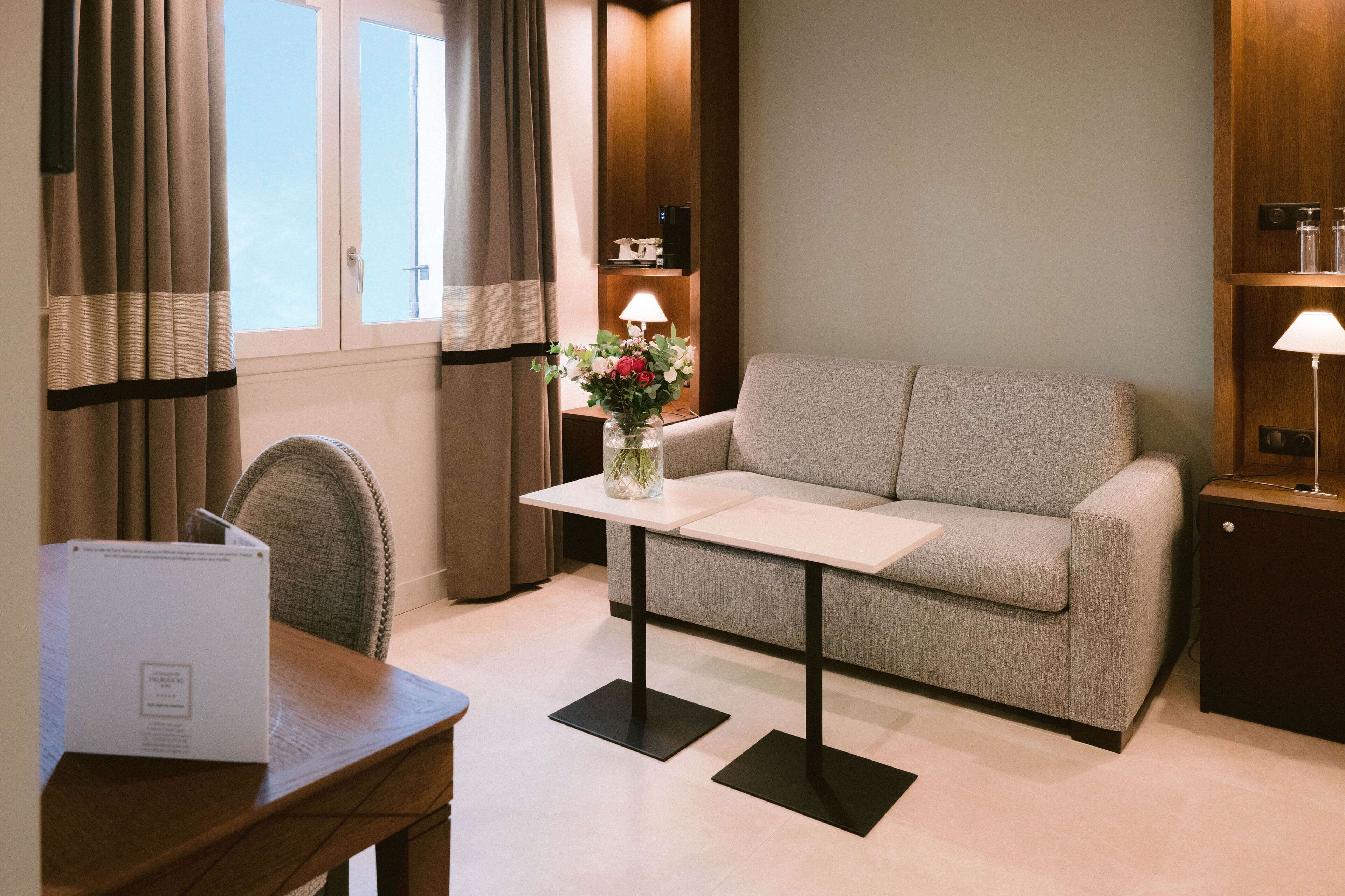 A modern hotel room with a grey sofa, a small table with a vase of flowers, a lamp, and a wooden nightstand.