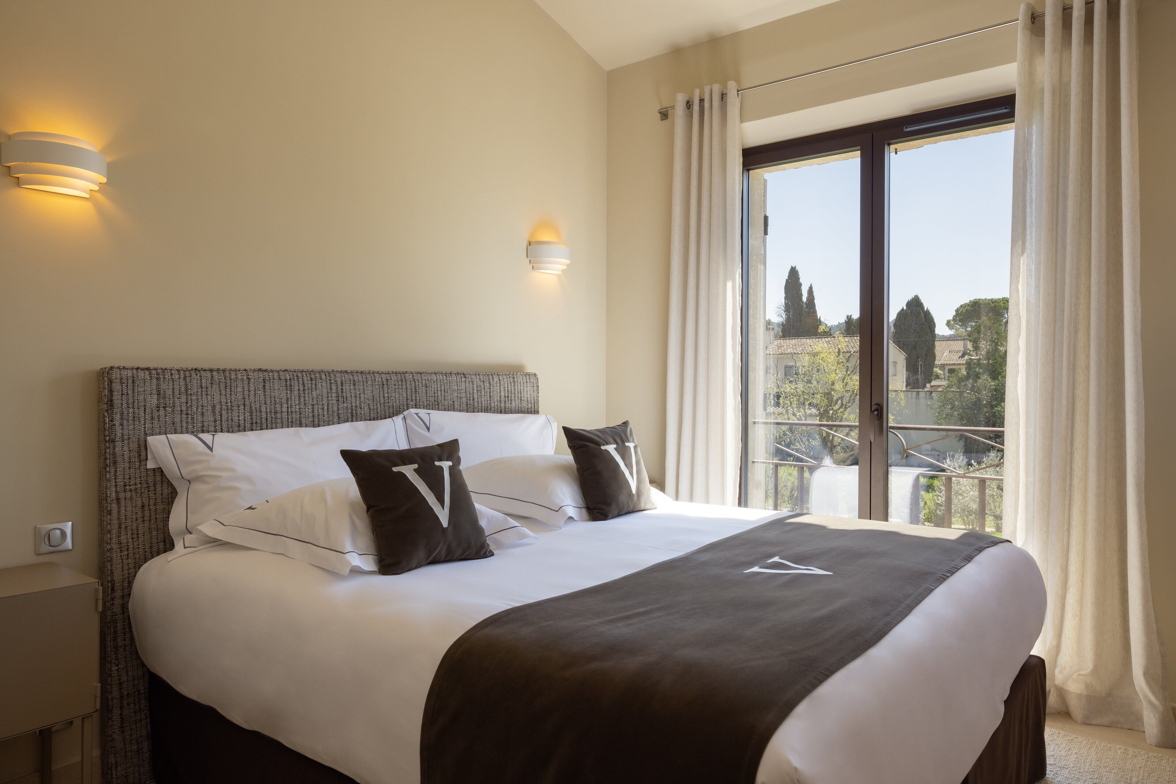 Ein Hotelzimmer mit einem großen Bett, zwei Nachttischlampen, Blick auf den Außenbalkon und einem mit Vorhängen versehenen Fenster.