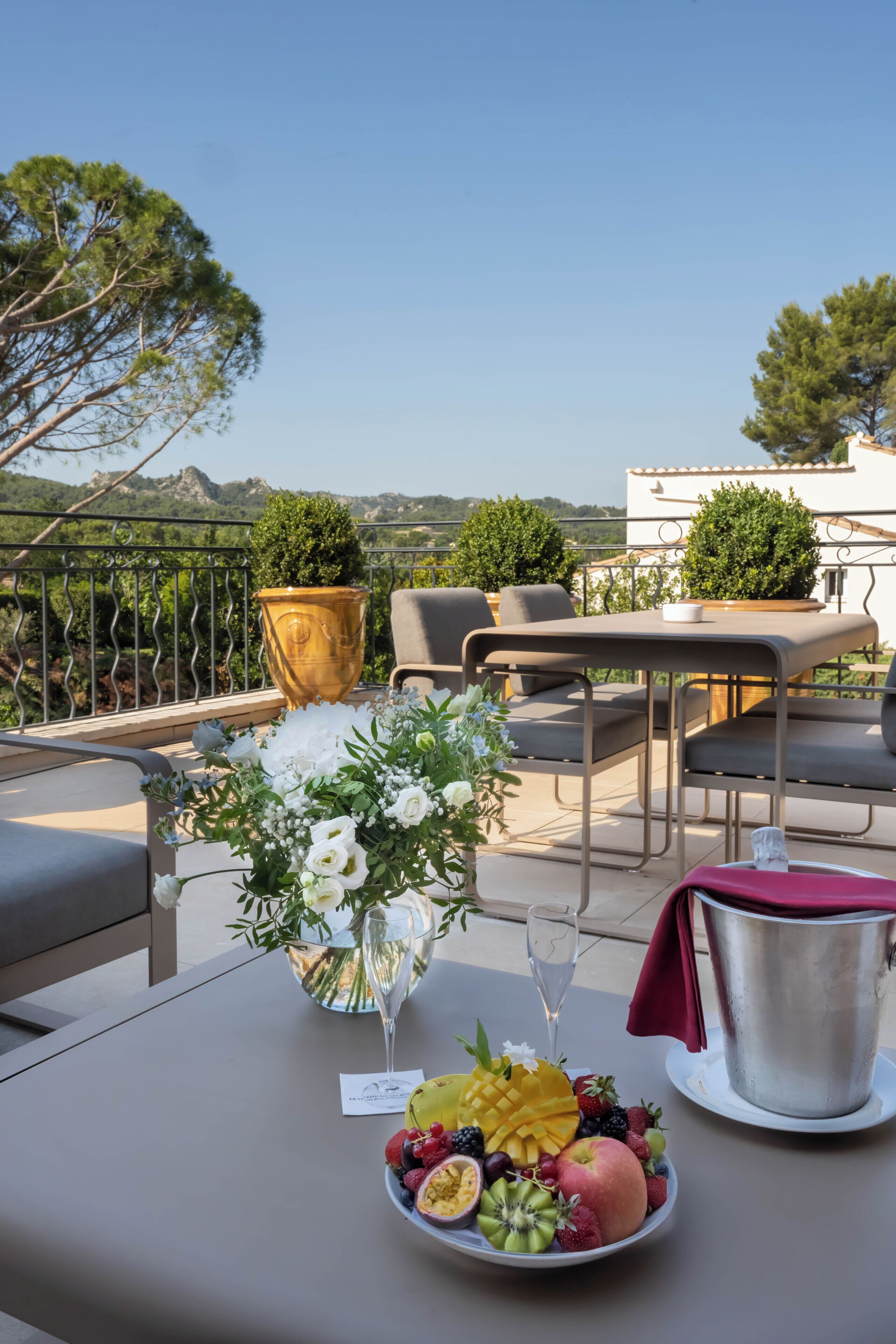 A terrace with a table set for a meal, a vase with flowers, and a fruit bowl.
