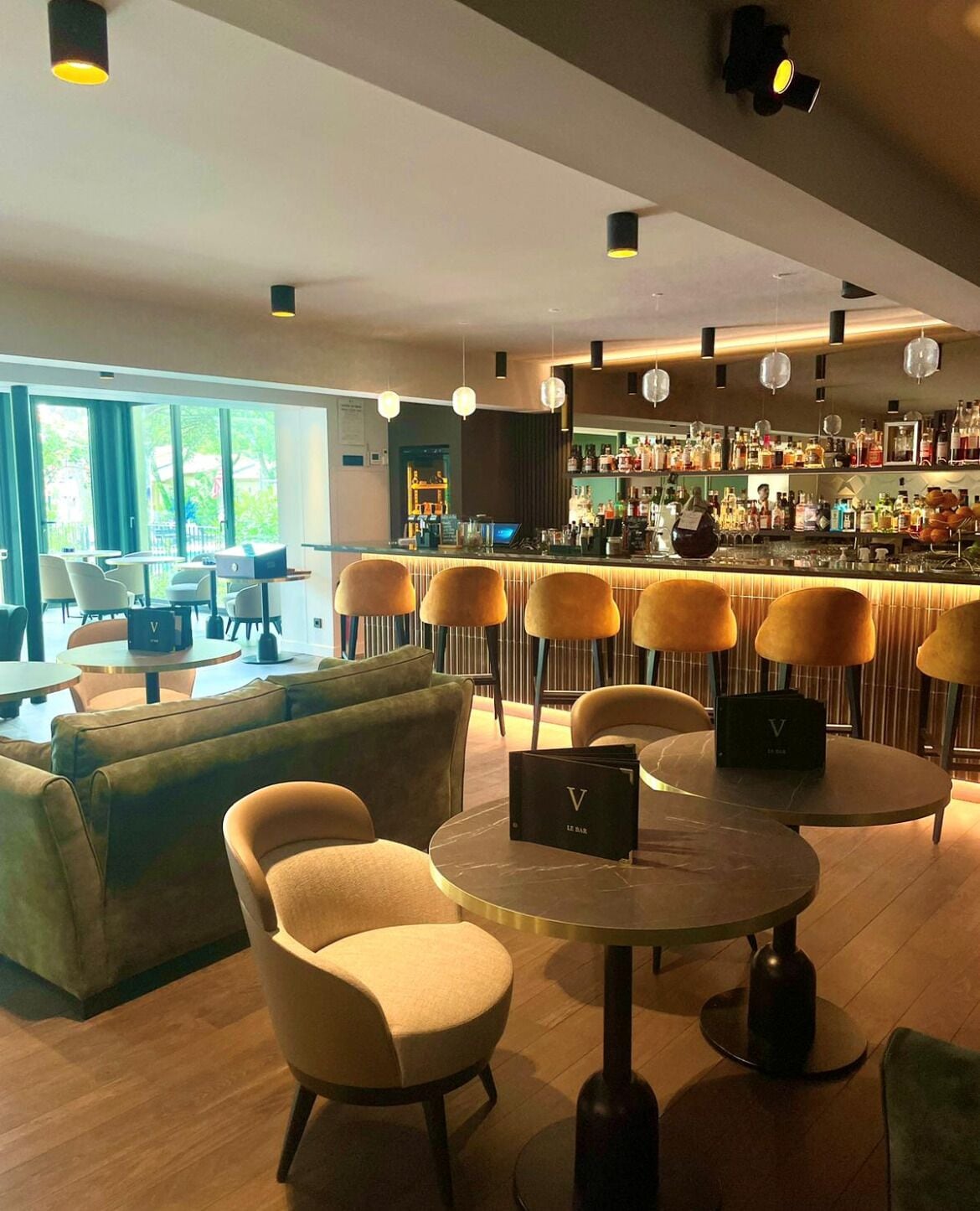 Un espace bar moderne avec des tables rondes et des tabourets, un coin salon avec des canapés et un comptoir de bar où sont exposées différentes bouteilles.