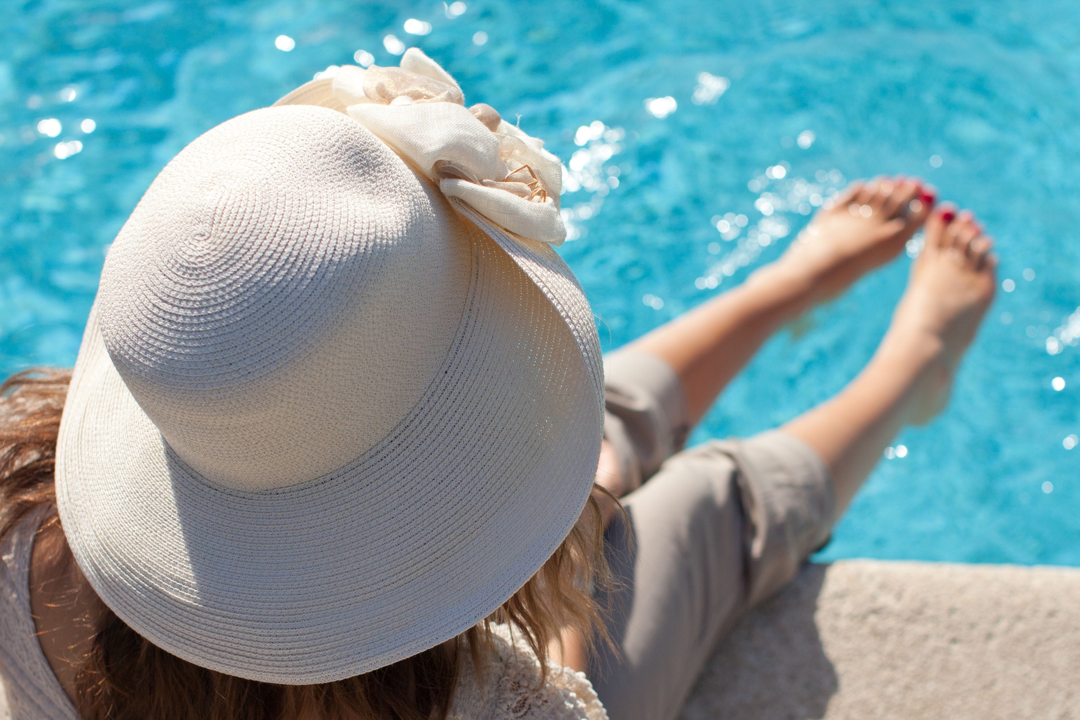 A person relaxing by a pool with a white hat and red nail polish.