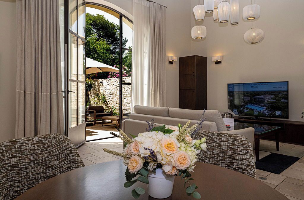 Ein modernes Wohnzimmer mit einer Couch, einem Couchtisch und einem Fernseher. Ein Esstisch mit floraler Dekoration steht ebenfalls im Raum. Große Fenster mit Vorhängen bieten einen Blick ins Freie.