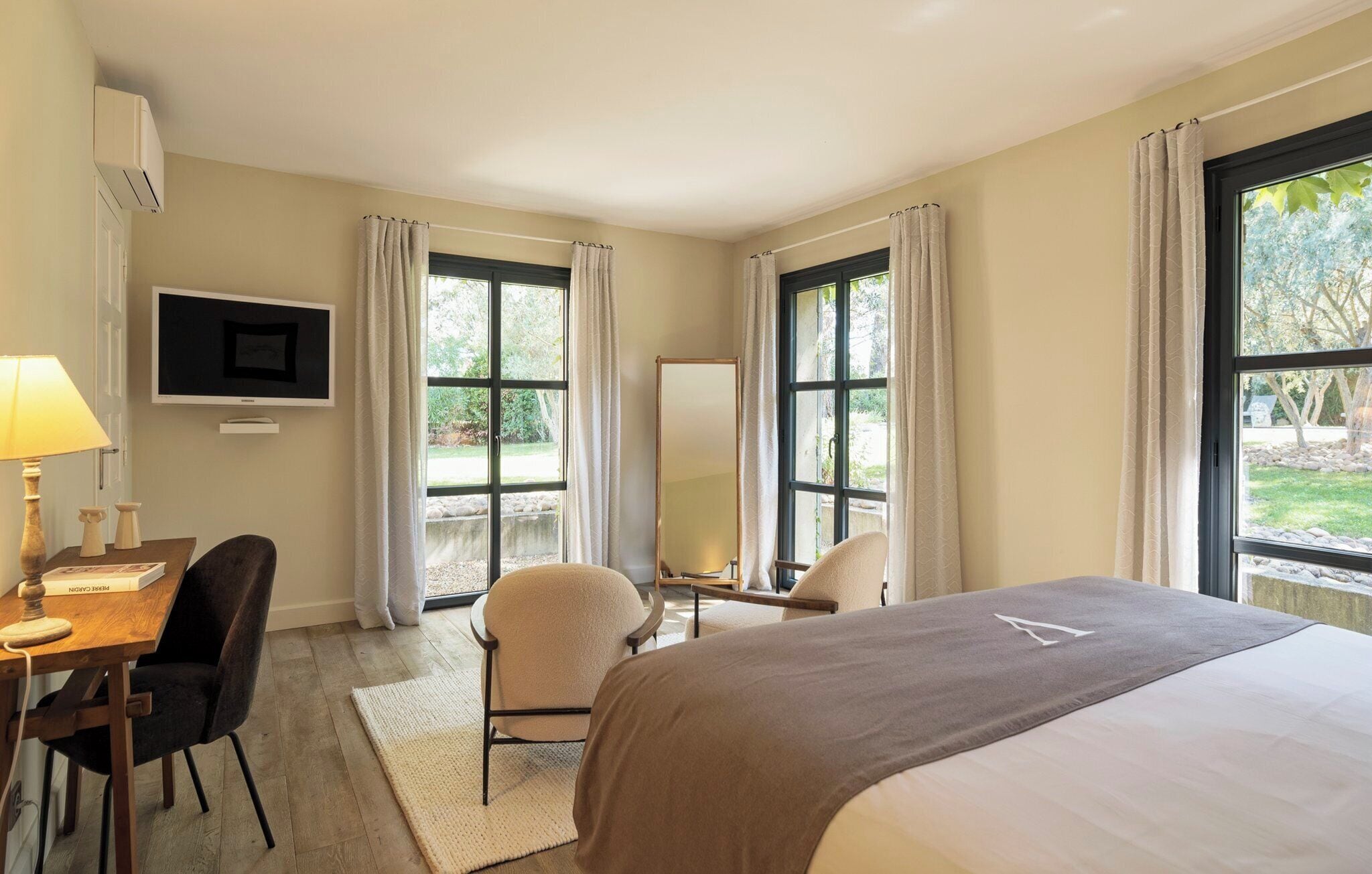 A hotel room with a bed, a desk with a lamp, a chair, a TV, and large windows with curtains.