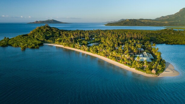 Vue aérienne d’un complexe hôtelier situé sur une île tropicale, avec une longue plage de sable, de nombreux palmiers et une eau d’un bleu cristallin.