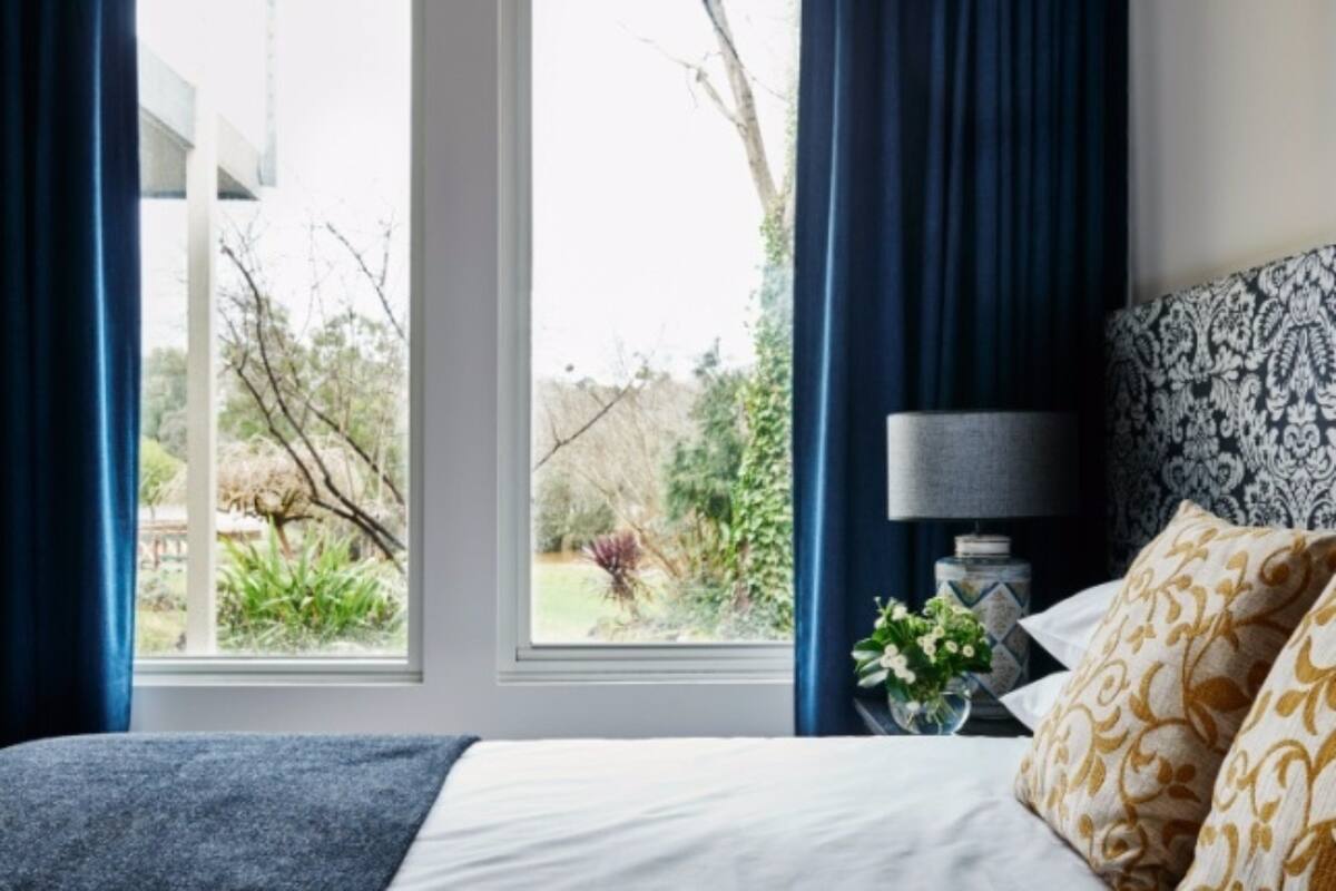 Une chambre avec une grande fenêtre, un lit agrémenté d’oreillers à motifs, une table de chevet avec une lampe, et une vue sur la verdure à l’extérieur.