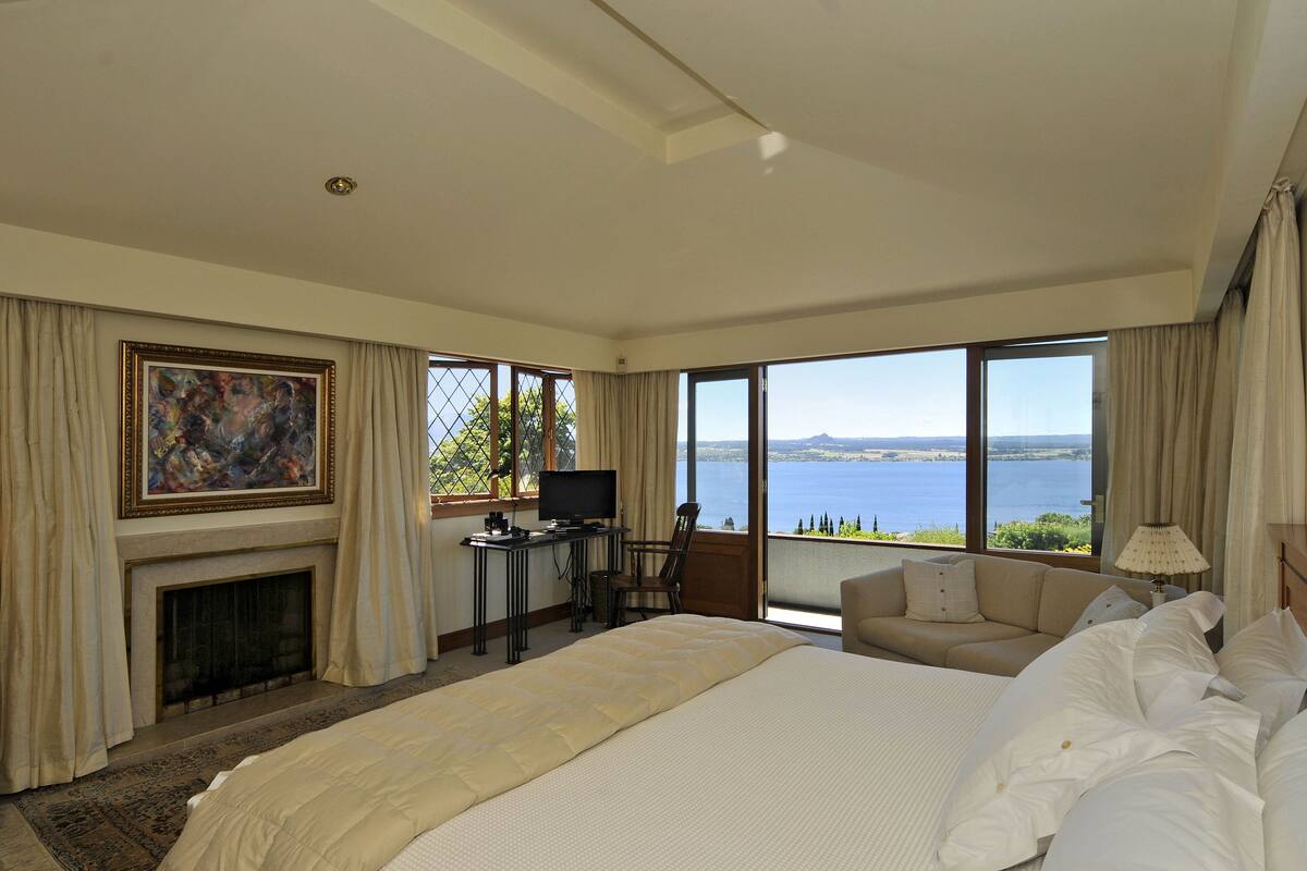 Une chambre d’hôtel avec un grand lit, un bureau avec une télévision, une cheminée, un canapé et une vue sur la mer.