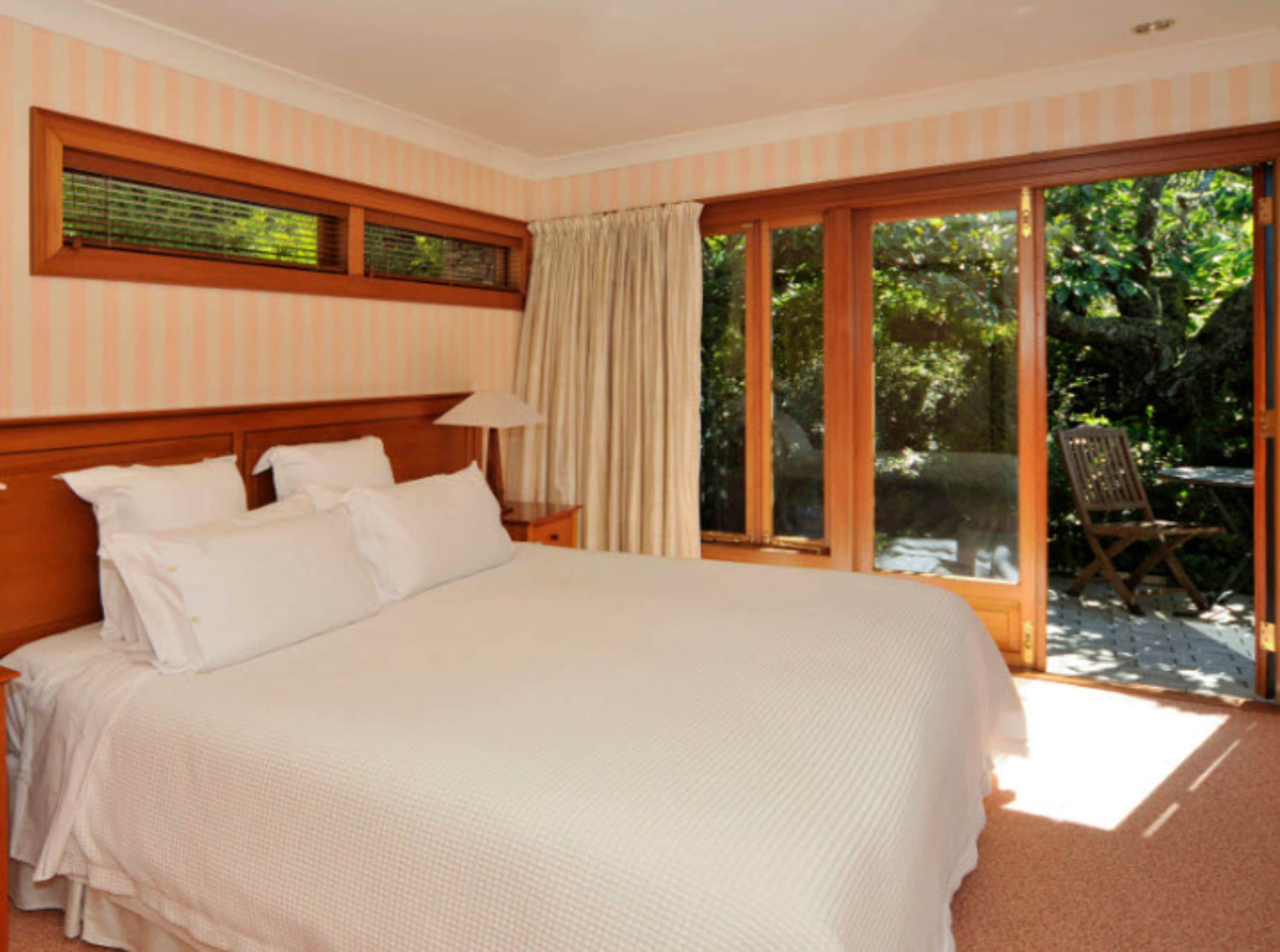 A bedroom with a large bed, a nightstand, a window with blinds, and a view of the outdoors.