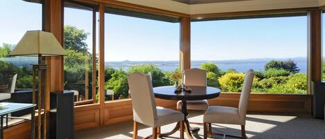 A corner room with a large window offering a view of greenery and a distant landscape. There is a round table with chairs, a lamp, and a speaker.