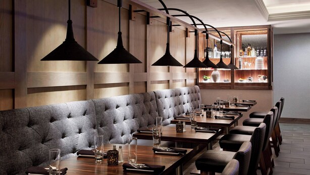 Ein Restaurant mit einer langen, gepolsterten Sitzbank, Holztischen und hängenden Pendelleuchten.