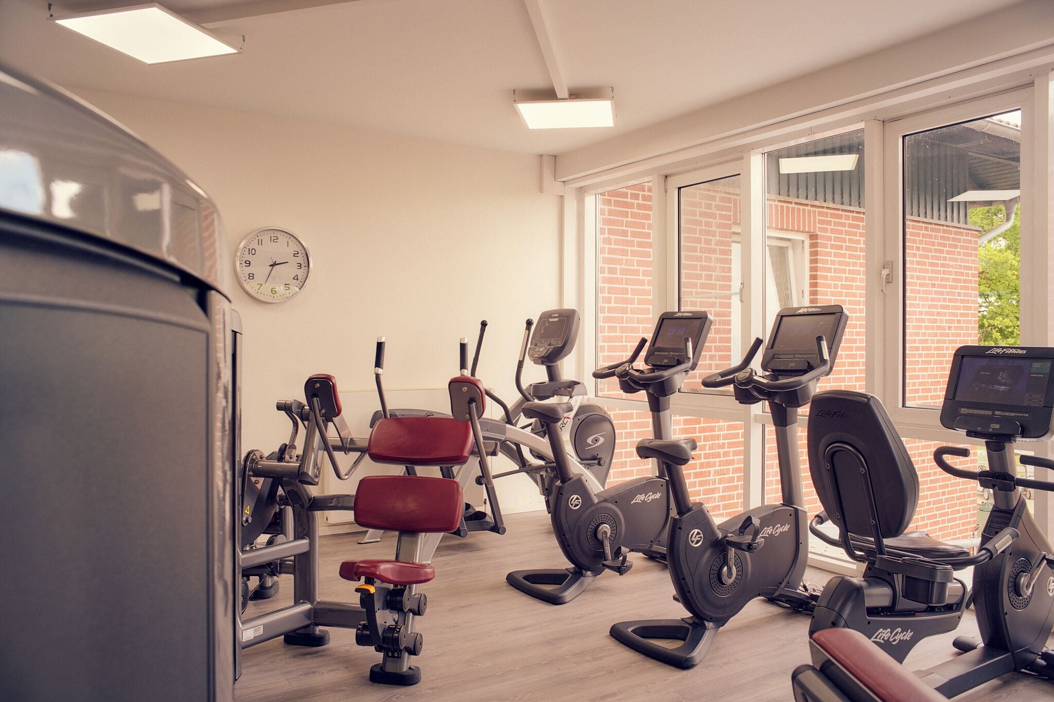 Een sportschool met diverse fitnessapparatuur, een klok aan de muur en grote ramen.
